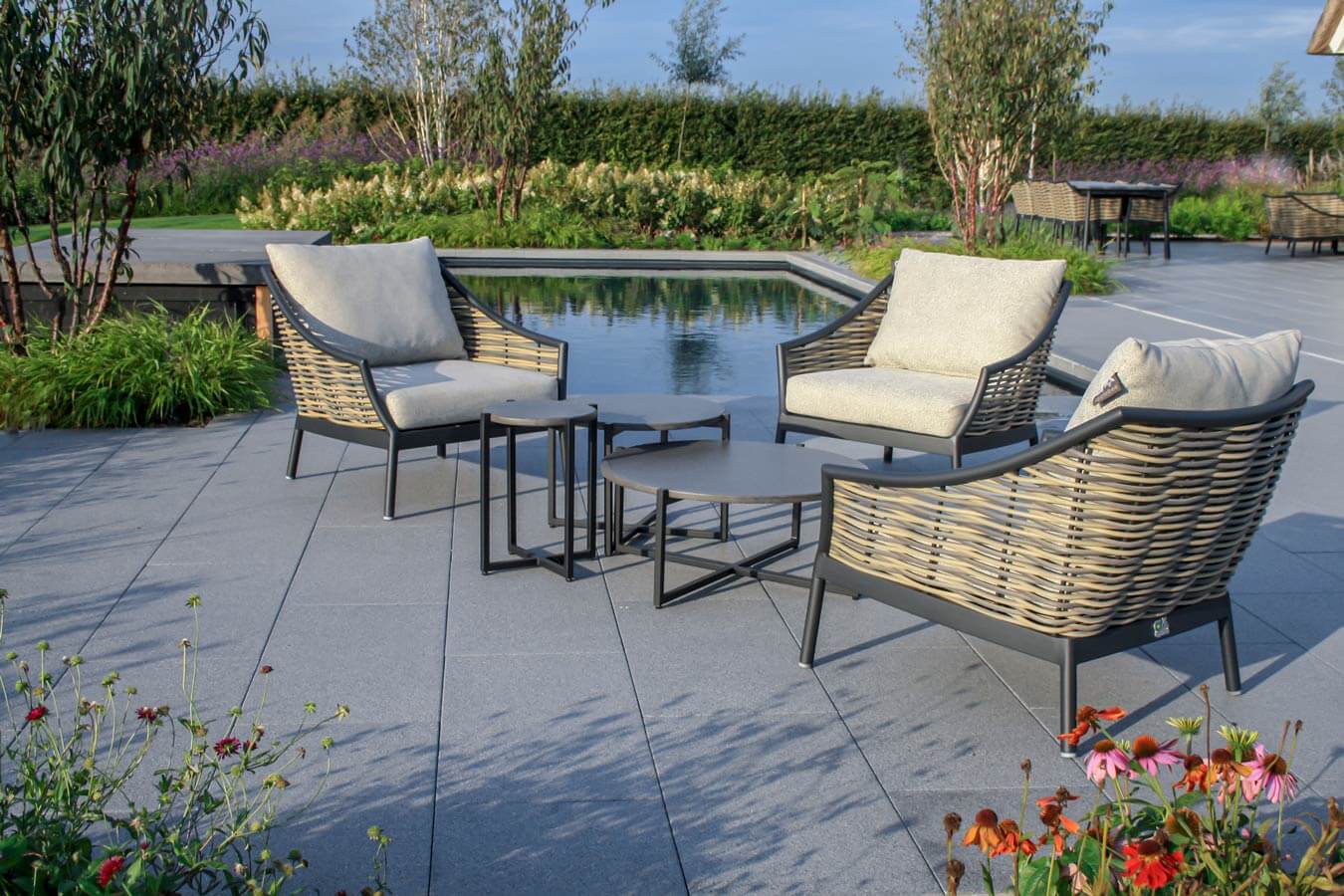 Outdoor seating area with stylish rattan chairs and tables by a pool, surrounded by greenery and flowers.