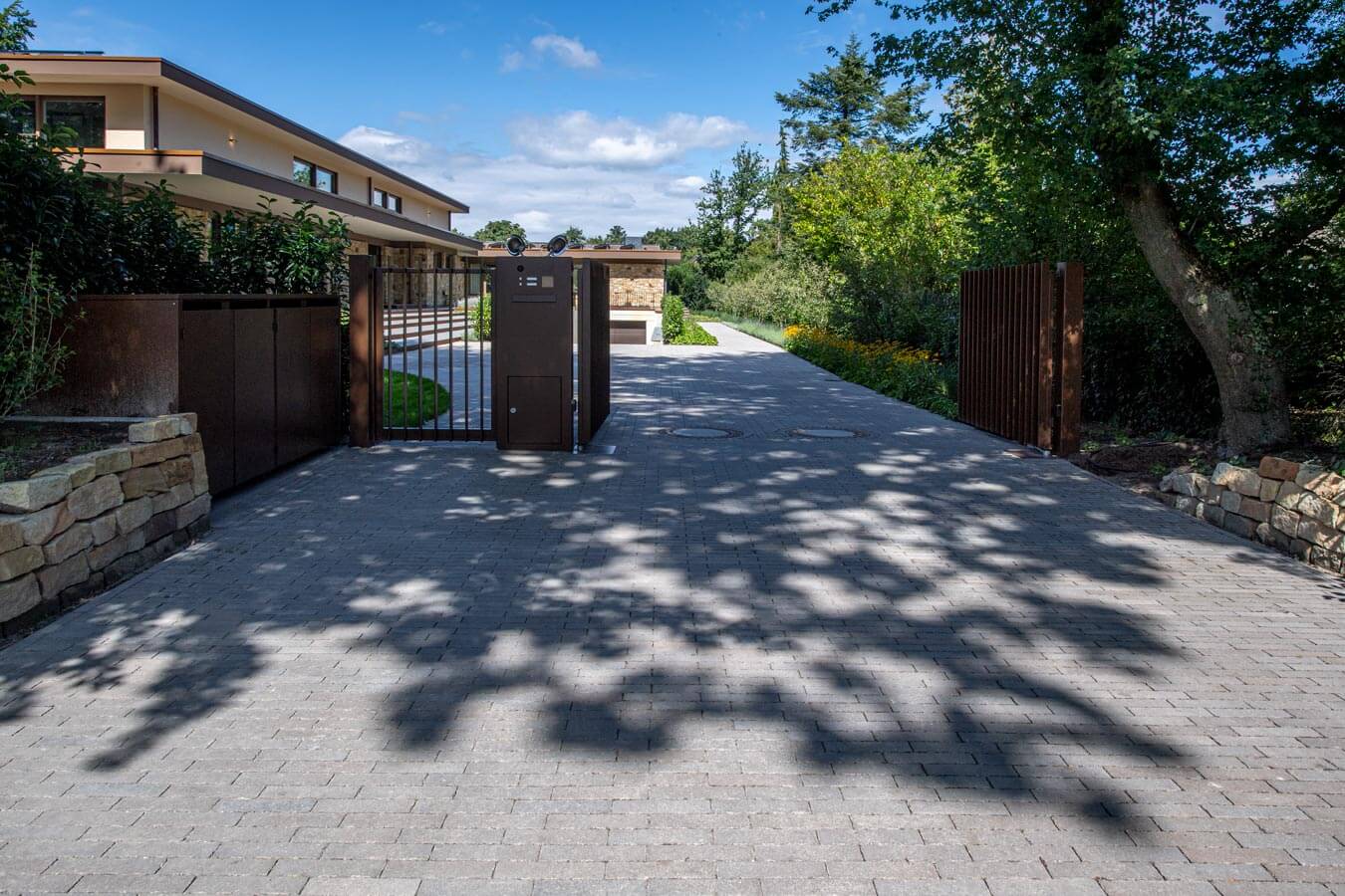 Pflastersteine METTEN SPRING ALVAR Braun-Grau auf einer Einfahrt mit Tor und umgebender Natur.