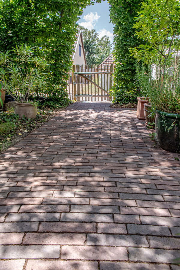 Pflastersteine METTEN SPRING ALVAR Rotschwarz auf einem lebendigen Gartenweg unter Schatten von Pflanzen.