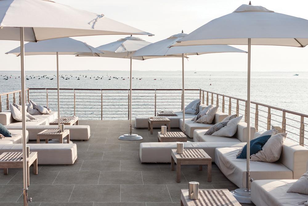 Eine stilvolle Terrasse am Wasser mit weißen Sonnenschirmen, bequemen Lounge-Sofas und Holztischen. Die Sitzgelegenheiten sind mit Kissen dekoriert, und im Hintergrund erstreckt sich ein ruhiges Gewässer mit kleinen Bojen und vereinzelten Booten. Die Terrasse hat einen modernen Metallgeländer-Abschluss und einen grauen Fliesenboden.